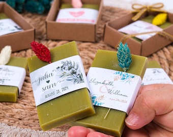 May include: Green rectangular soaps with decorative floral accents and personalized labels. The labels feature names and wedding dates. Each soap is topped with a dried flower and presented in a small brown box, ideal for wedding favors.