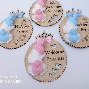 Op de afbeelding: Houten babyshower-cadeautjes met een wit babybeeldje dat een roze of blauwe muts en een kroon draagt. De cadeautjes zijn versierd met vlinders en de tekst "Welkom Prinses" of "Welkom Prins".