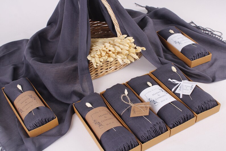 May include: Six grey scarves in brown boxes with white labels that say "Marry & Edward 2023" and a small dried flower sprig on top of each scarf.
