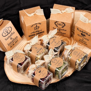 Op de afbeelding: Een verzameling gepersonaliseerde huwelijksbedankjes, waaronder kleine vierkante glazen potjes gevuld met thee en kleine bruine dozen met linten. De dozen en potjes zijn voorzien van namen en data, perfect voor een bruidsdouche of bruiloft.