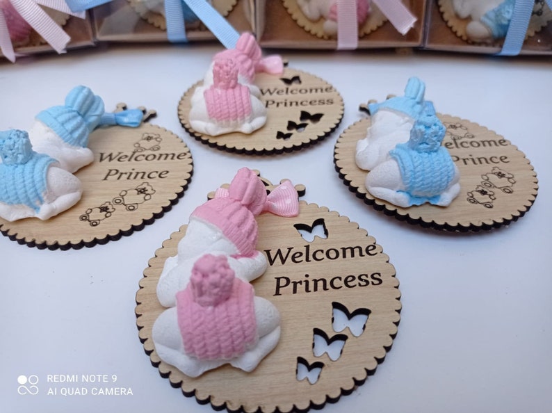 Op de afbeelding: Twee houten baby shower-cadeautjes met een slapende babyfiguur op elk. Een cadeautje is blauw en zegt "Welkom Prins" en het andere is roze en zegt "Welkom Prinses".
