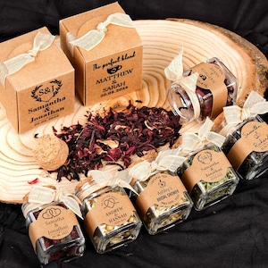 Puede incluir: Una colección de recuerdos de boda personalizados con pequeños frascos de vidrio y cajas de papel kraft. Los frascos y cajas están llenos de hojas de té y hierbas, y adornados con etiquetas y cintas personalizadas. Los recuerdos están dispuestos sobre una rodaja de madera.