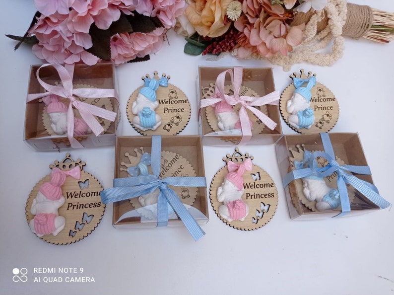 Op de afbeelding: Acht kleine geschenkdozen met doorzichtige deksels, elk met een houten cirkel met een babyfiguur en de woorden "Welkom Prins" of "Welkom Prinses". De dozen zijn gebonden met blauw of roze lint.