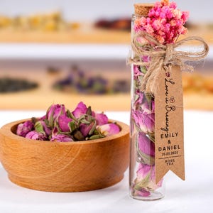 Può includere: Una ciotola di legno piena di boccioli di rosa rosa secchi è accanto a una fiala di vetro piena di boccioli di rosa e sormontata da fiori rosa. La fiala è legata con spago e un'etichetta con "Love is Brewing" e i nomi "Emily & Daniel" con una data.