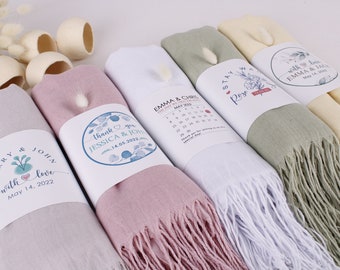 May include: Four different wedding favour scarves with personalised labels. The scarves are in shades of grey, pink, white, and green. The labels feature floral designs and wedding dates.