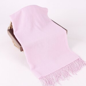 May include: A soft, light pink scarf with a fringe border. The scarf is draped over a brown wicker basket.