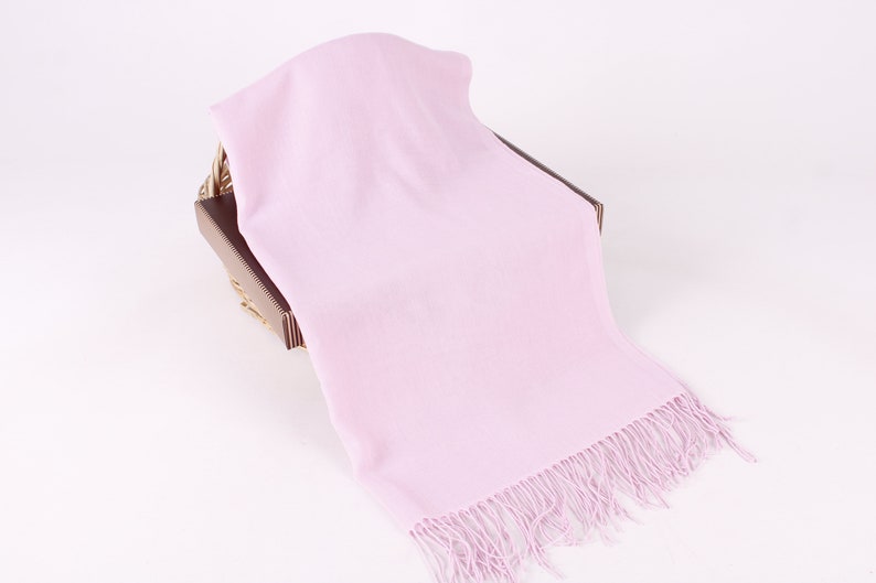 May include: A soft, light pink scarf with a fringe border. The scarf is draped over a brown wicker basket.