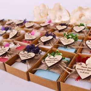 Può includere: Una collezione di piccole saponette decorative in scatole di cartone individuali. Ogni saponetta è avvolta con spago e sormontata da fiori secchi e un'etichetta di legno a forma di cuore con la scritta "Thank you Kenroy & Sherine 12.18.2021".
