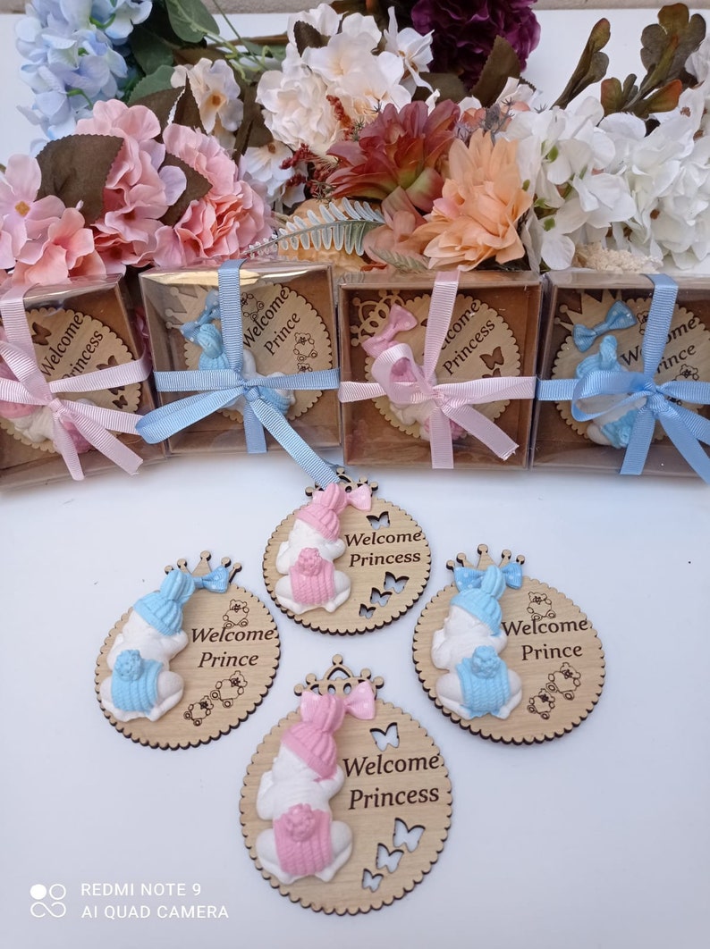Op de afbeelding: Houten babyshower-cadeautjes met een wit babybeeldje dat een roze of blauwe muts en een kroon draagt. De cadeautjes zijn versierd met vlinders en de tekst "Welkom Prinses" of "Welkom Prins".