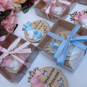 Op de afbeelding: Een set babyshower-cadeautjes met een klein, wit, slapende babybeeldje met een roze of blauwe muts. Het beeldje is bevestigd aan een houten cirkel met de tekst "Welkom Prinsesje" of "Welkom Prins" en een kroonontwerp. De cadeautjes zijn verpakt in doorzichtige doosjes met een roze of blauw lint.