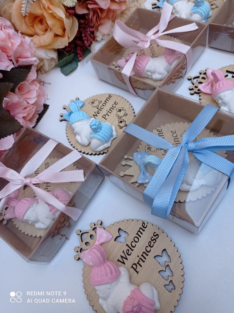 Op de afbeelding: Een set babyshower-cadeautjes met een klein, wit, slapende babybeeldje met een roze of blauwe muts. Het beeldje is bevestigd aan een houten cirkel met de tekst "Welkom Prinsesje" of "Welkom Prins" en een kroonontwerp. De cadeautjes zijn verpakt in doorzichtige doosjes met een roze of blauw lint.