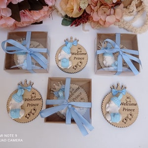Op de afbeelding: Vier kleine houten cirkels met een babyfiguurtje dat een blauwe muts en een kroon draagt. Elke cirkel heeft de tekst "Welcome Prince" erop. De cirkels zijn vastgebonden met een blauw lint en zitten in kleine doosjes.