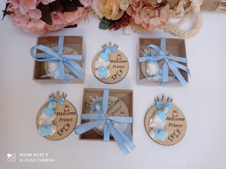 Op de afbeelding: Vier kleine houten cirkels met een babyfiguurtje dat een blauwe muts en een kroon draagt. Elke cirkel heeft de tekst "Welcome Prince" erop. De cirkels zijn vastgebonden met een blauw lint en zitten in kleine doosjes.