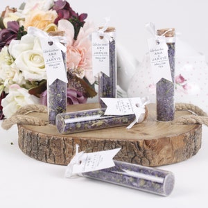 May include: Glass tubes filled with mallow tea, each with a cork stopper and a white tag. The tags read "Ana & Jarvis Mallow Tea." The tubes are arranged on a wooden slice with a bouquet of flowers in the background.