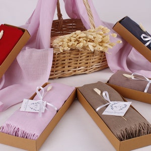 May include: Five gift boxes containing different coloured scarves. The boxes are brown and tied with white ribbon. Each box has a small white tag with a handwritten message. The scarves are red, pink, grey, brown and beige.