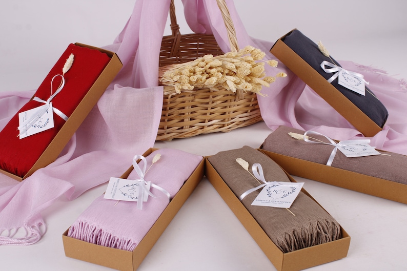 May include: Five gift boxes containing different coloured scarves. The boxes are brown and tied with white ribbon. Each box has a small white tag with a handwritten message. The scarves are red, pink, grey, brown and beige.