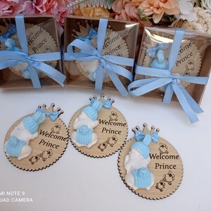 Op de afbeelding: Vier ronde houten ornamenten met een blauw en wit babybeeldje en de tekst "Welcome Prince". De ornamenten zitten in kleine doorzichtige doosjes die zijn vastgebonden met een blauw lint.