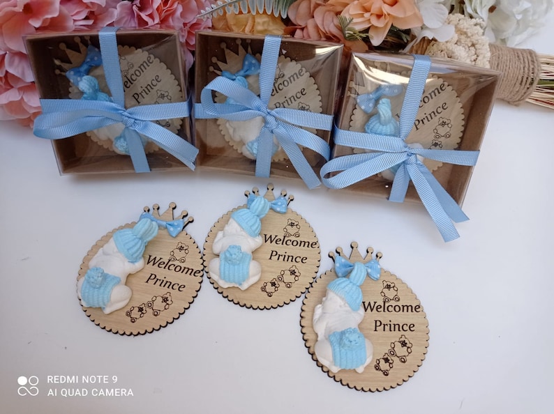 Op de afbeelding: Vier ronde houten ornamenten met een blauw en wit babybeeldje en de tekst "Welcome Prince". De ornamenten zitten in kleine doorzichtige doosjes die zijn vastgebonden met een blauw lint.
