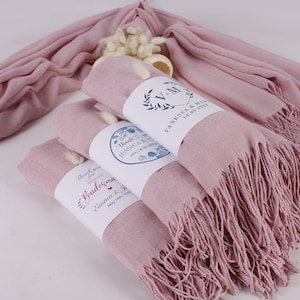 May include: Stack of four pink pashmina scarves with white fringe. Each scarf is wrapped with a white label with blue and red text. The labels read "Thank you for being a Bridesmaid", "Thank you for being a Bridesmaid", "Thank you for being a Bridesmaid", and "Vanessa & Mike 14.05.2023".