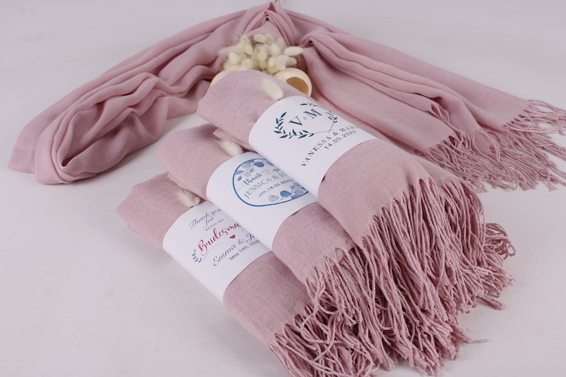 May include: Stack of four pink pashmina scarves with white fringe. Each scarf is wrapped with a white label with blue and red text. The labels read "Thank you for being a Bridesmaid", "Thank you for being a Bridesmaid", "Thank you for being a Bridesmaid", and "Vanessa & Mike 14.05.2023".