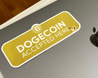 May include: A gold and white sticker on a gray laptop. The sticker reads "DOGECOIN ACCEPTED HERE" with the Dogecoin logo and a cartoon Shiba Inu dog. The laptop has the Apple logo.