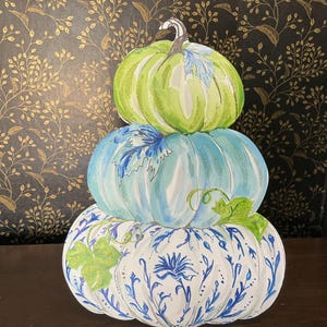 May include: A decorative cutout featuring three stacked pumpkins. The top pumpkin is light green, the middle is light blue with a blue leaf, and the bottom is white with blue floral patterns and green leaves. The design has a watercolor aesthetic.