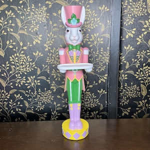 May include: A whimsical Easter bunny figurine in a pink and green outfit, holding a white tray. The bunny wears a pink top hat with a blue flower and a green bow tie. The base is yellow with purple diamonds.
