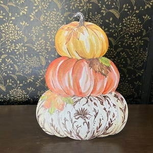 May include: A decorative cutout featuring a stack of three pumpkins. The top pumpkin is yellow, the middle is orange with a brown leaf, and the bottom is white with intricate brown floral line art and a small orange leaf accent on the side.