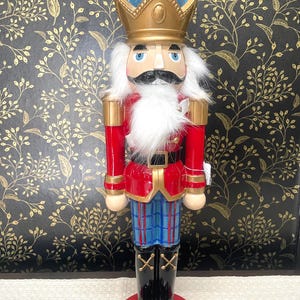 May include: A decorative nutcracker figurine. The figurine is dressed in a red jacket with gold accents, blue and red striped pants, and black boots. It has a white beard and mustache, and a blue and gold crown. The base is red.