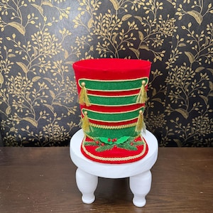 May include: A cylindrical red and green felt hat featuring gold braided trim, two horizontal green bands, and gold tassels. The base is decorated with green pine needles and red berries, all resting on a small white pedestal with four legs.
