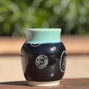 May include: A small, handmade ceramic vase with a black body and a turquoise rim. The vase features white, spiral-like designs. The base is white, and the vase is sitting on a wooden surface. The background is blurred green foliage.