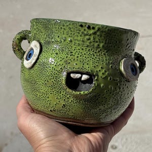 May include: A handmade, green ceramic bowl with a textured surface and a whimsical monster face design. The bowl features two blue-eyed eyeballs, a toothy grin, and two small handles. The bowl is held in a hand.