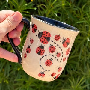 May include: A ceramic mug with a cream-colored exterior featuring a repeating pattern of red and black ladybugs. The mug has a glossy black handle and a matching black interior. A dashed black line curves across the surface, tracing the flight path of the ladybugs.
