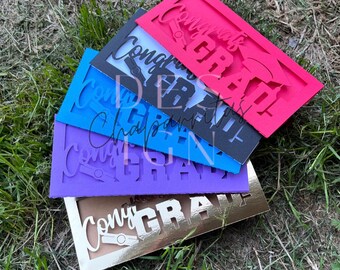 May include: A stack of colorful graduation cards in pink, black, blue, purple, and gold. Each card features the words "Congrats Grad" cut out in a decorative font, along with a graduation cap illustration. The cards are arranged on a bed of green grass.