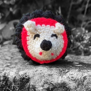 Pu&ograve; includere: Un animal di peluche all'uncinetto a forma di riccio con un viso rosso e bianco e occhi neri. Il riccio &egrave; seduto su una superficie di pietra grigia.