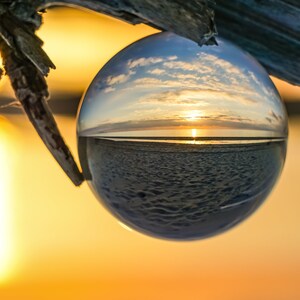 Könnte beinhalten: Eine Glaskugel fängt einen malerischen Sonnenuntergang über einem Gewässer ein, mit einem bewölkten Himmel und einem Sandstrand im Vordergrund.