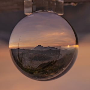 Può includere: Una sfera di cristallo che riflette una catena montuosa al tramonto. Le montagne sono in silhouette contro un cielo rosa e arancione. La sfera di cristallo è appesa a un soffitto.