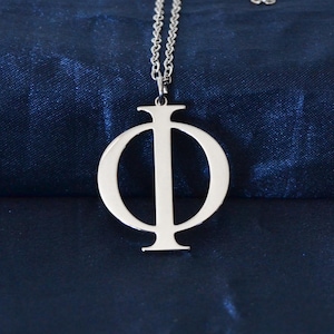 Può includere: Una collana con pendente color argento raffigurante la lettera greca Phi (Φ). Il pendente è appeso a una delicata catena su uno sfondo blu scuro strutturato. La collana è un gioiello semplice ed elegante.