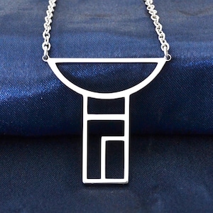 May include: A silver-colored pendant necklace with a geometric design. The pendant features a half-circle above a rectangular shape with cut-out geometric details. The necklace chain is also silver and rests on a dark blue surface.