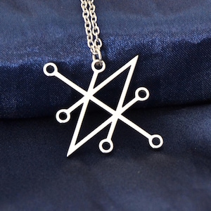Op de afbeelding: Zilverkleurige hangerketting met een geometrisch ontwerp van lijnen en cirkels. De hanger hangt aan een delicate ketting tegen een donkerblauwe stoffen achtergrond. De ketting is een sieraad.