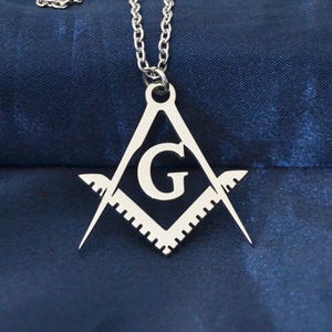 Peut inclure: Collier argenté avec un pendentif représentant un symbole maçonnique. Le pendentif est en forme d'équerre et de compas avec la lettre "G" au centre. Le collier est posé sur un tissu bleu foncé satiné.