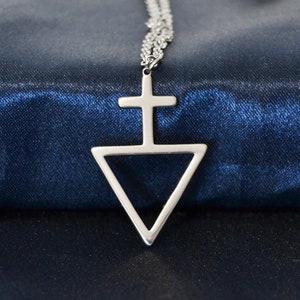 Può includere: Collana con pendente color argento raffigurante un simbolo stilizzato: una croce sopra un triangolo capovolto. La catena è sottile e il pendente è posto su uno sfondo blu scuro strutturato. Un gioiello.