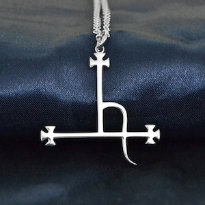 Può includere: Una collana con un ciondolo d'argento che presenta una croce stilizzata con una linea curva che si estende dalla parte inferiore. Il ciondolo è su uno sfondo di tessuto blu scuro.