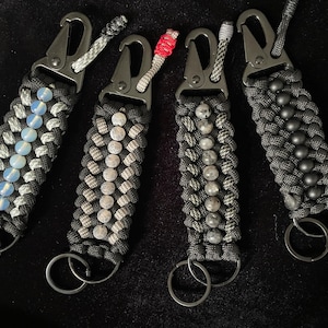 May include: Five black paracord keychains with different colored beads. The keychains are made with black paracord and have a silver metal clasp. The beads are made of different materials, including glass, stone, and metal.