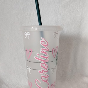 May include: A clear plastic tumbler with a green straw. The tumbler is decorated with pink hearts, white bows, and the name "Caroline" in pink script. The Starbucks logo is visible.