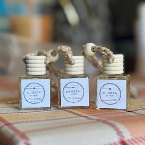 Puede incluir: Tres pequeñas botellas cuadradas de vidrio con tapas de madera y colgantes de cuerda. Cada botella tiene una etiqueta que dice "Blueberry Cobbles". Las botellas están dispuestas sobre una superficie a cuadros.