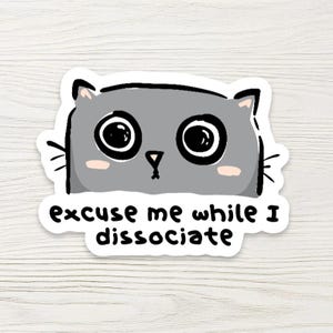 May include: A die-cut sticker featuring a gray cartoon cat with large black eyes, a small pink nose, and light pink cheeks. The cat has black whiskers and a black outline. Below the cat, the text reads: excuse me while I dissociate.