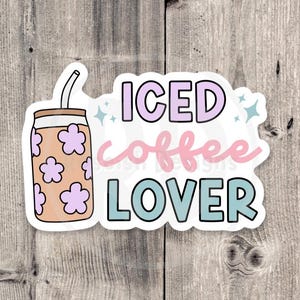 Puede incluir: Una pegatina con las palabras "ICED coffee LOVER" en colores pastel. El dise&ntilde;o incluye un caf&eacute; helado de dibujos animados con detalles florales. El fondo es una superficie de madera.