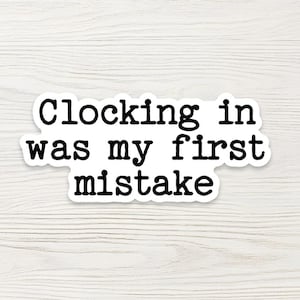 May include: A white sticker with the text "Clocking in was my first mistake" in a bold, black font. The sticker has a rounded edge and is set against a light wood grain background.
