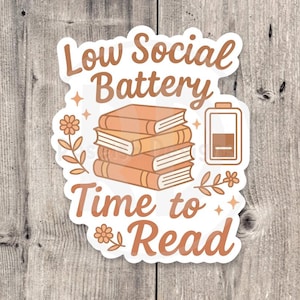 May include: A sticker with the text "Low Social Battery Time to Read" in brown script. The design includes a stack of books, a battery icon, flowers, and stars, all in shades of brown and orange.
